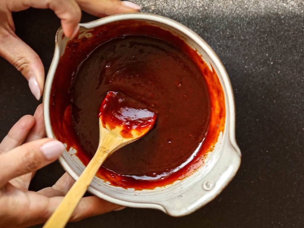 Hands hold a bowl of dark red gochujang sauce, with a small wooden spoon resting inside. The background is a dark textured surface.